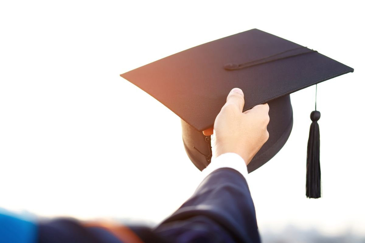 Close-up of a person wearing a graduation gown, holding a black graduation cap with a tassel against a bright background, contemplating how to choose a college.