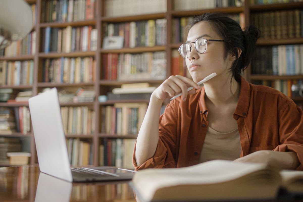 A young Asian woman with glasses pondering while looking away from her laptop in a library setting, with books and a lamp beside her.