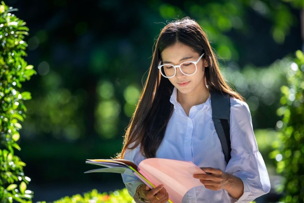 A young woman in glasses reads a book outdoors, sunlight filtering through trees in the background.