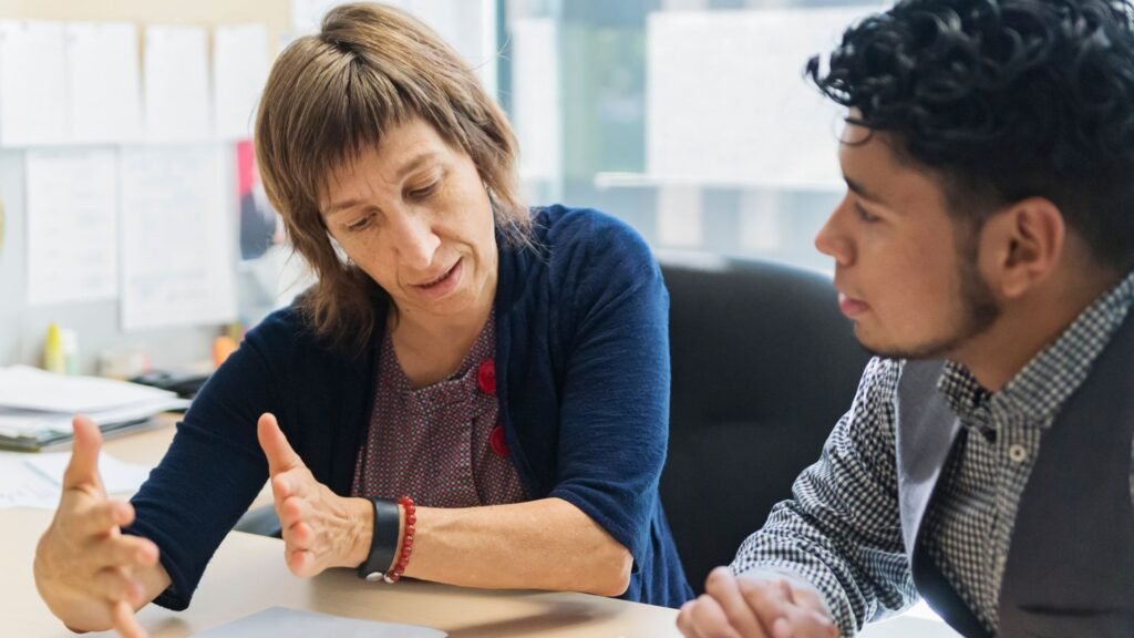 Two people sitting at a desk, engaged in a conversation. One person gestures with their hands while the other listens attentively. Office supplies and paper notices are visible in the background.