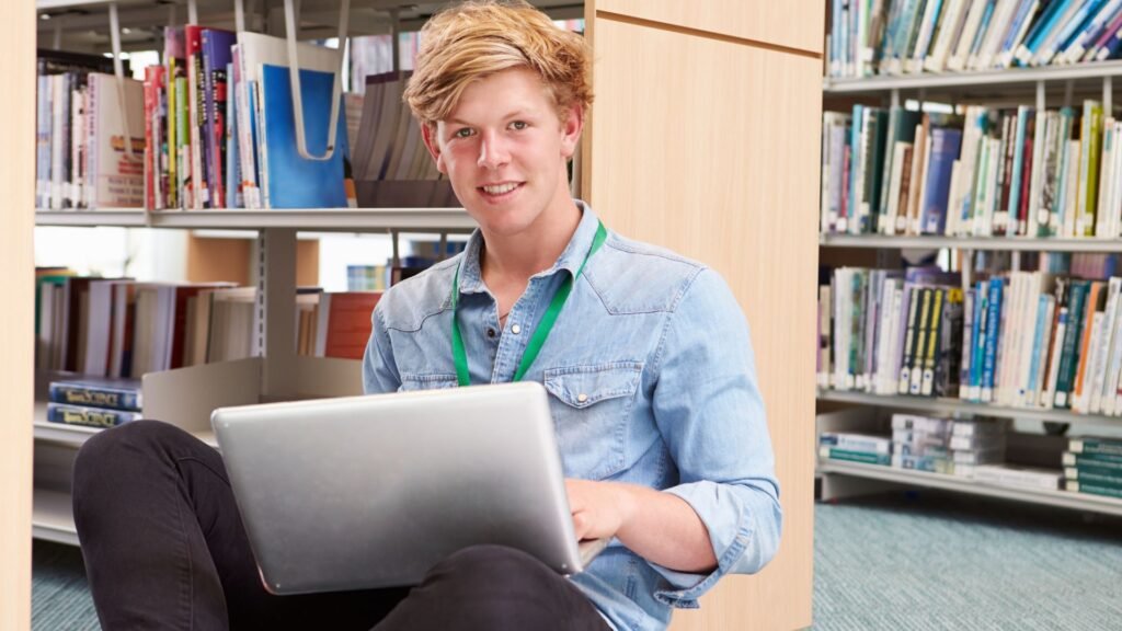 A young person sits on the floor of a library, working on a laptop and smiling at the camera, a Senior Year Checklist visible on the screen, with bookshelves in the background.