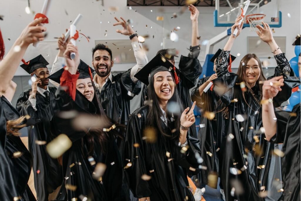 Graduates in caps and gowns cheer and celebrate, holding diplomas, with confetti falling around them in a gymnasium.