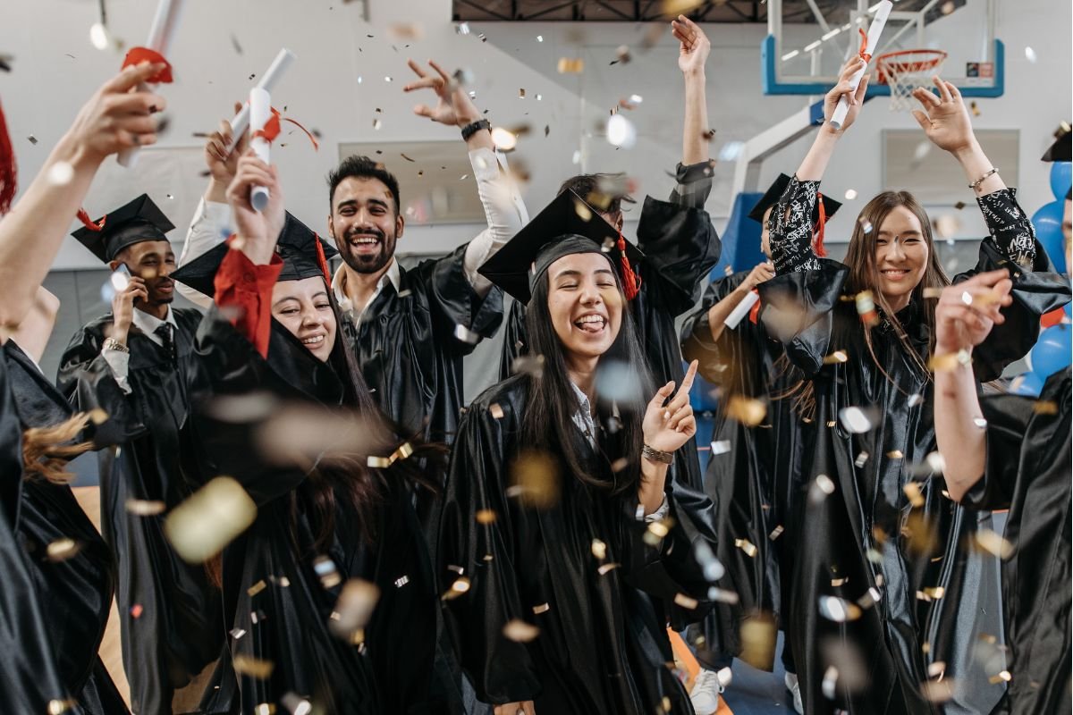 Graduates in caps and gowns cheer and celebrate, holding diplomas, with confetti falling around them in a gymnasium.