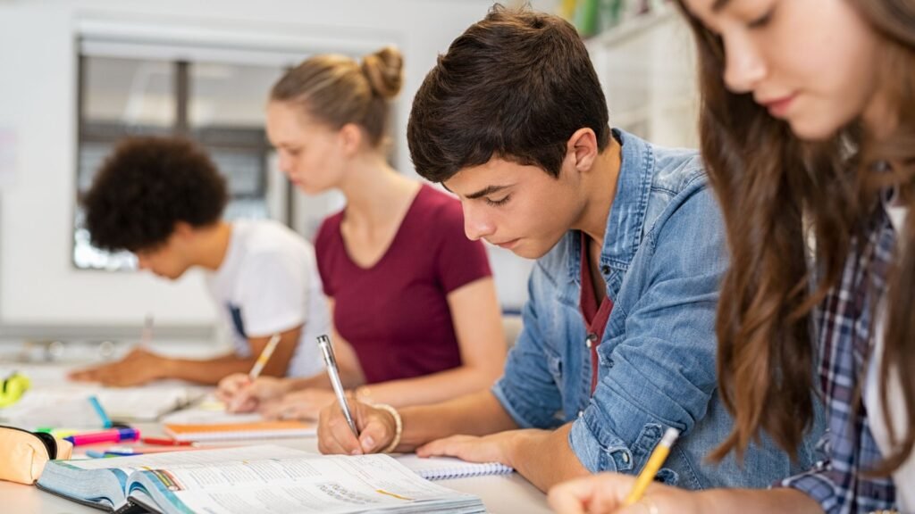 Four students sit in a classroom, writing in notebooks and using textbooks. They appear focused on their work, pondering questions like "Does High School GPA Matter After High School?" Pens, notebooks, and other school supplies are scattered on the table.