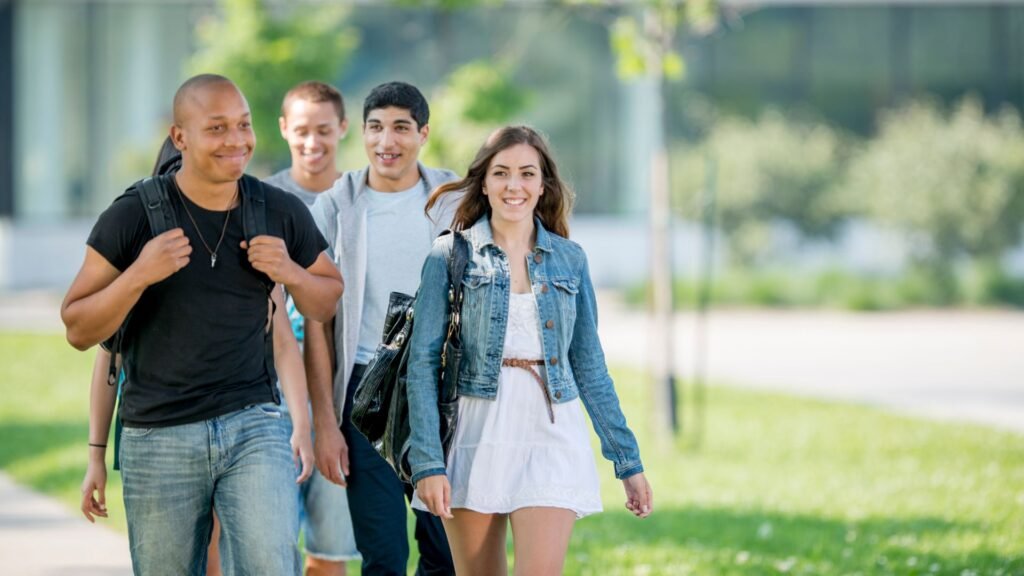 A group of four young adults, three men and one woman, walking outdoors on a sunny day. They appear to be students, carrying backpacks and smiling, perhaps discussing their Senior Year Checklist.