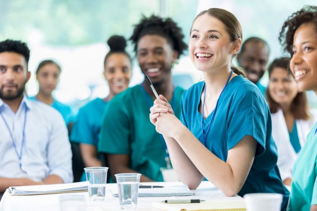 A group of healthcare professionals in scrubs attend a meeting. A woman in the foreground, smiling and holding a pen, sits at a table with notebooks and glasses of water.