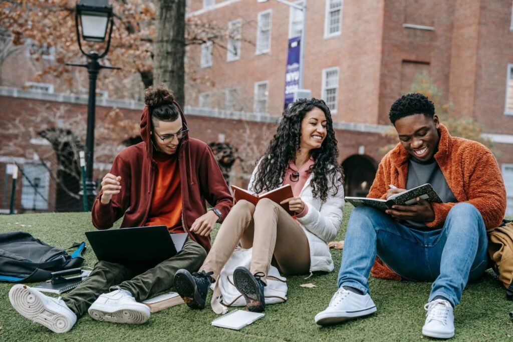 Three people sit on grass reading and using a laptop. Buildings and a lamp post are in the background. They appear to be enjoying their time together.