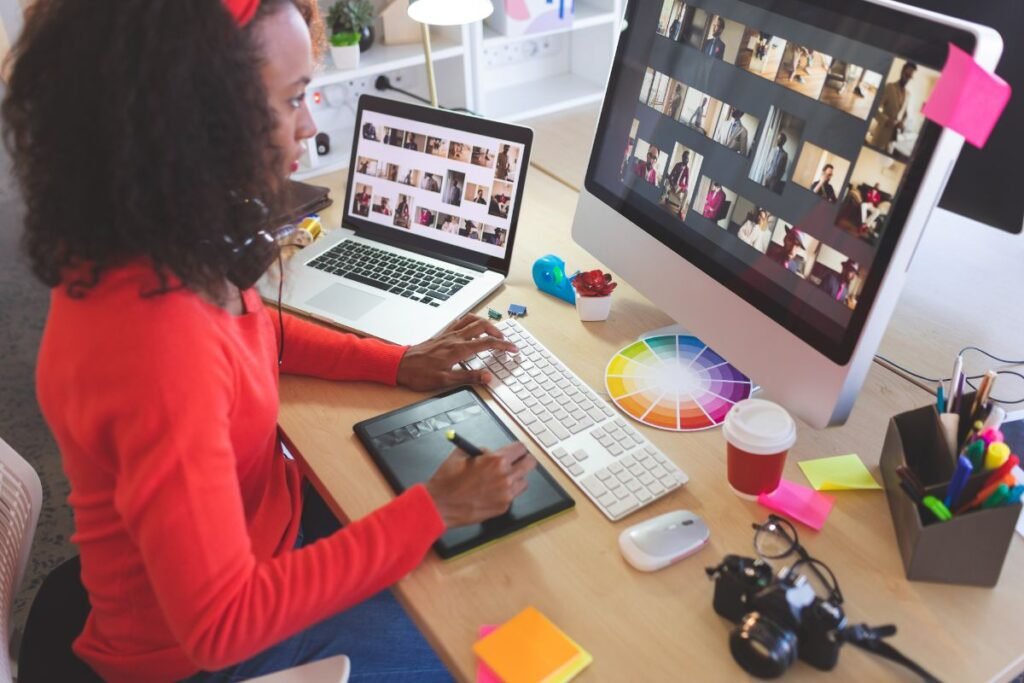 A woman is using a tablet and computer for photo editing at a cluttered desk with a laptop, camera, coffee cup, and color wheel.