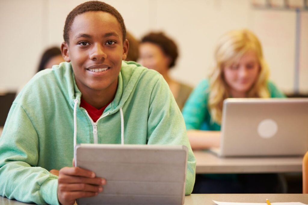 A student in a green hoodie holds a tablet, with classmates using laptops in the background.