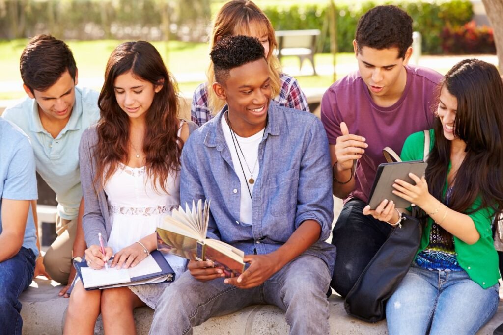 A group of diverse young adults sitting outdoors, engaging with books and a tablet.