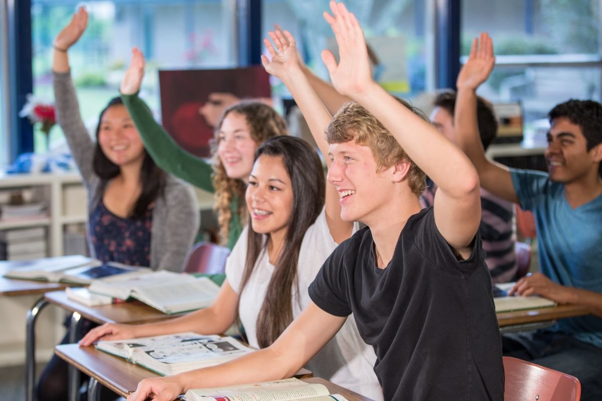 Students sitting at desks in a classroom, raising their hands, with textbooks open in front of them.