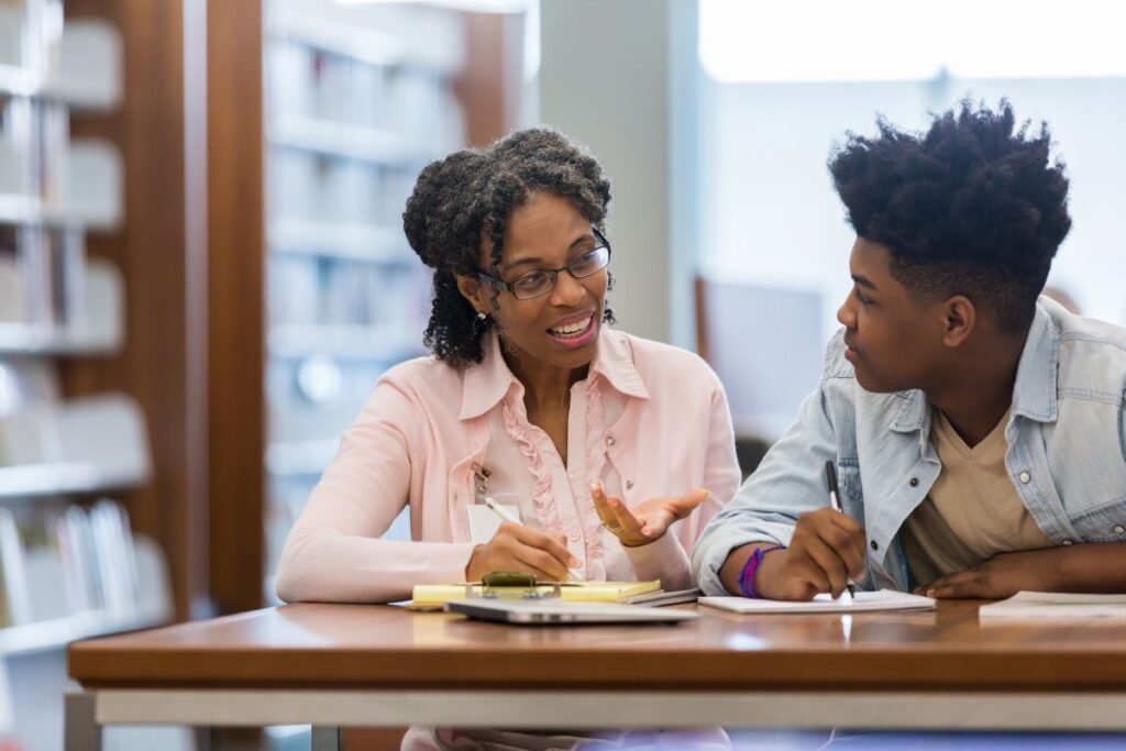 A teacher and student sit at a library table, discussing notes. Both have notebooks and pens, with bookshelves in the background.