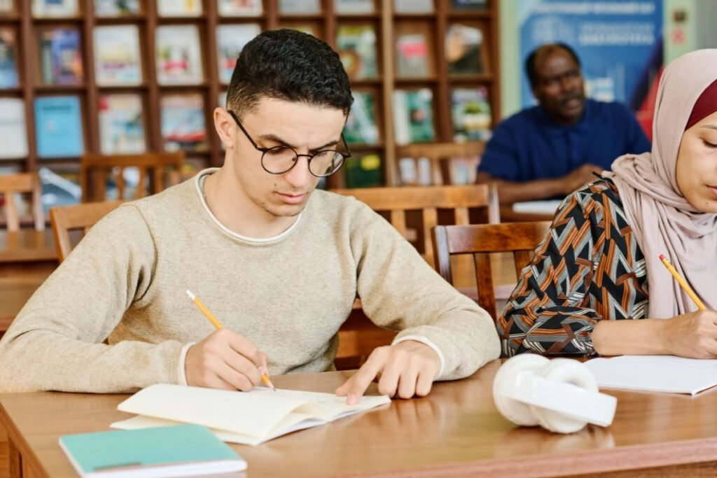 A man in glasses studies, writing in a notebook, with a woman beside him. A man is visible in the background. They're seated at a wooden table in a library.