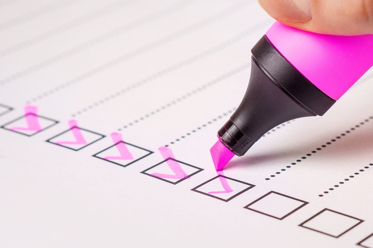 A hand uses a pink highlighter to check off boxes on a white checklist with black lines and text.