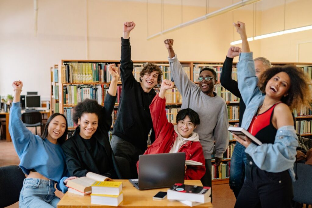 A group of people in a library celebrate with raised fists, surrounded by open books and a laptop on the table, perhaps having just mastered the difference between AP and IB classes in high school.