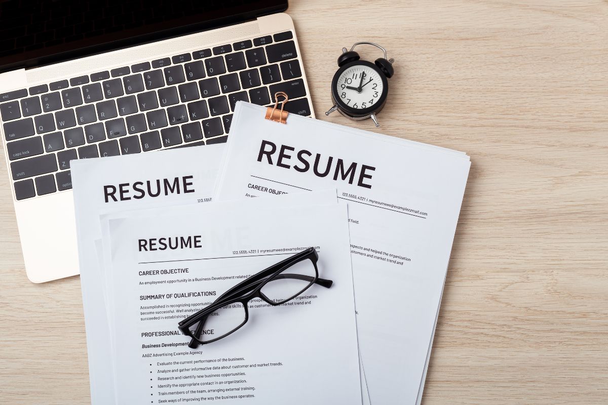 A stack of resumes for college applications rests on a desk, accompanied by a pair of glasses, a small clock, and an open laptop.