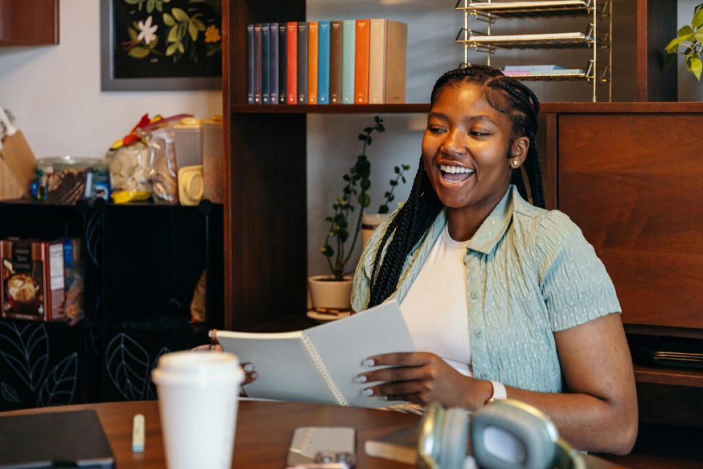 A person sits at a table, smiling and holding an open notebook that compares AP vs. IB classes in high school. A coffee cup and headphones are on the table, while shelves with books and plants provide a cozy backdrop.