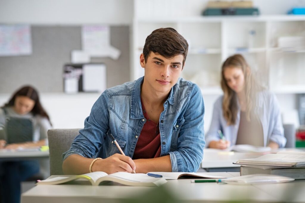 A teenage boy sits in a classroom, jotting down notes in his notebook, while two classmates engage in animated discussion about the difference between AP and IB classes in high school.