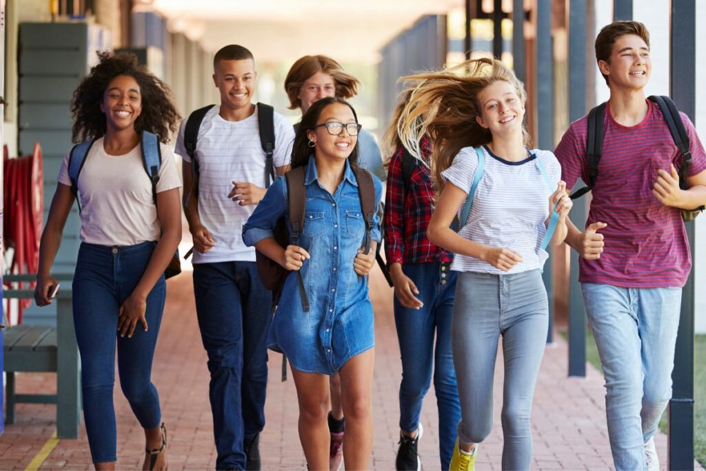 A group of six students, chatting about the difference between AP and IB classes in high school, walk together on a campus walkway, smiling in their casual clothes with backpacks.