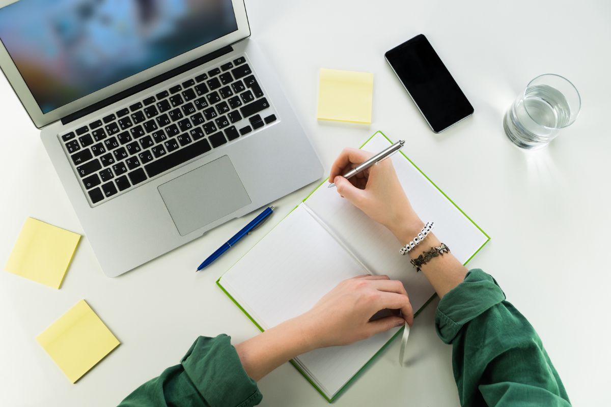 A person is diligently writing a college application resume in a notebook at a desk equipped with a laptop, smartphone, sticky notes, a pen, and a glass of water.