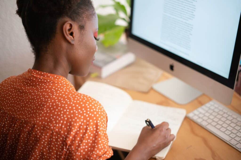 A person in an orange polka dot shirt writes in a notebook at a desk with an Apple computer, focusing intently on crafting the perfect college application resume.