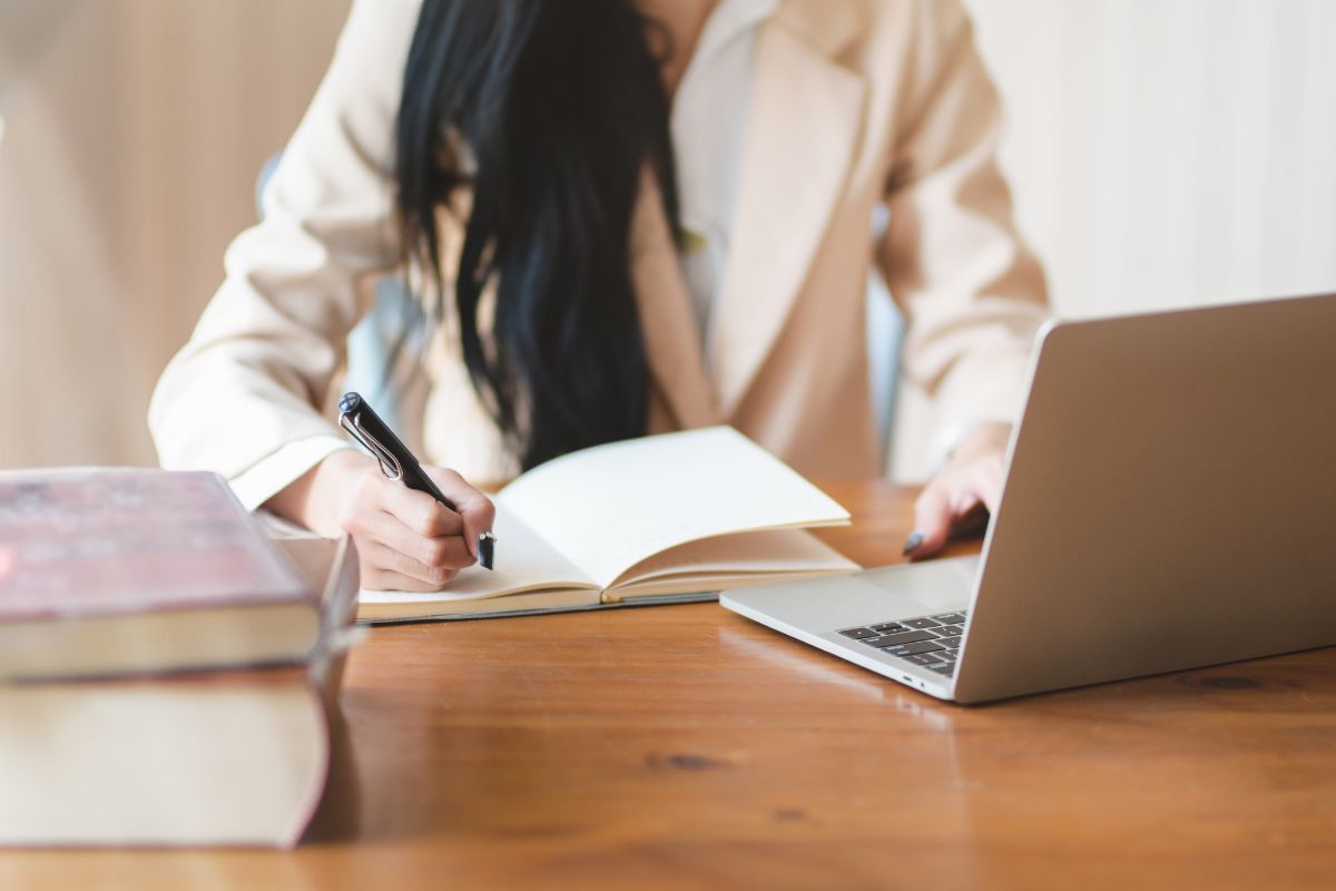 A person crafting a college application resume in a notebook beside their laptop on a wooden desk.