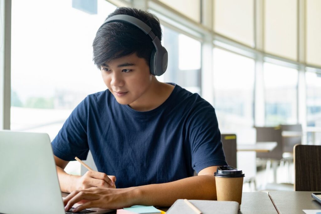 A person wearing headphones works on a laptop in a bright room with large windows, holding a pen. As if drafting the perfect college application resume, they sip coffee from a cup on the table.