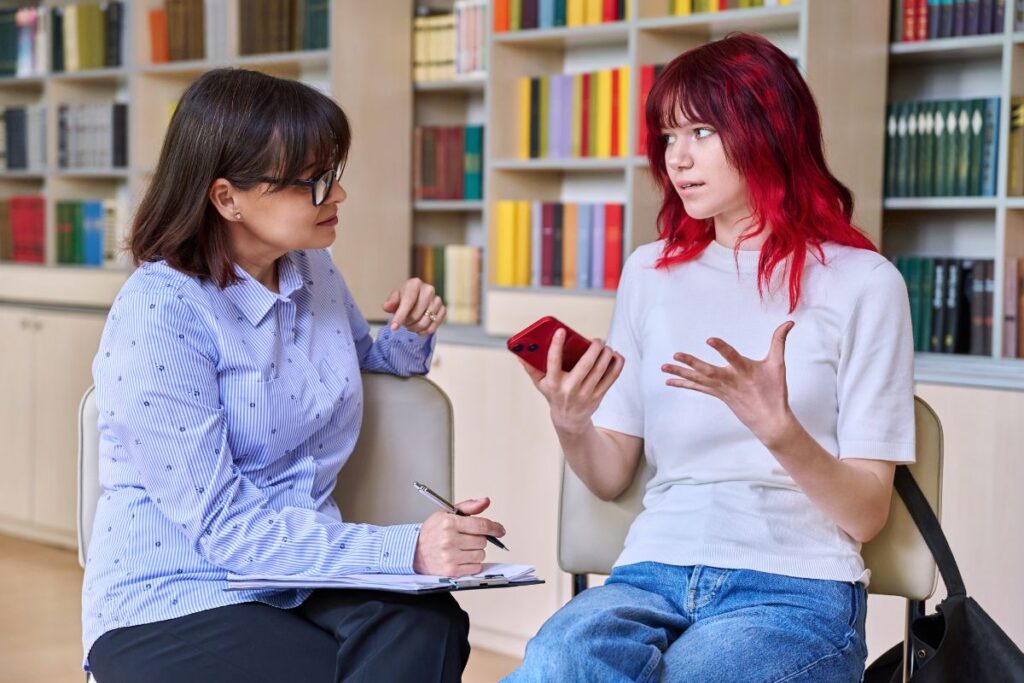 Two women sit in a library talking. One with red hair gestures while holding a phone, the other with glasses takes notes. Shelves with books are in the background.