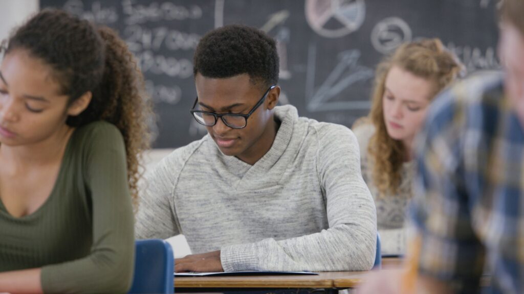 Students sitting at desks, focused on their work in a classroom with a chalkboard in the background, ponder if submitting SAT scores is necessary for college admissions or if Ivy League schools remain test-optional.