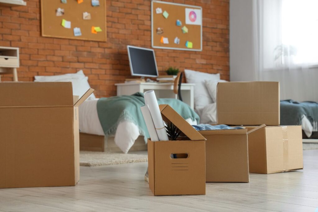 Room with cardboard boxes on the floor, a bed, desk, and corkboards on a brick wall. Boxes contain packed items, suggesting moving or organizing.