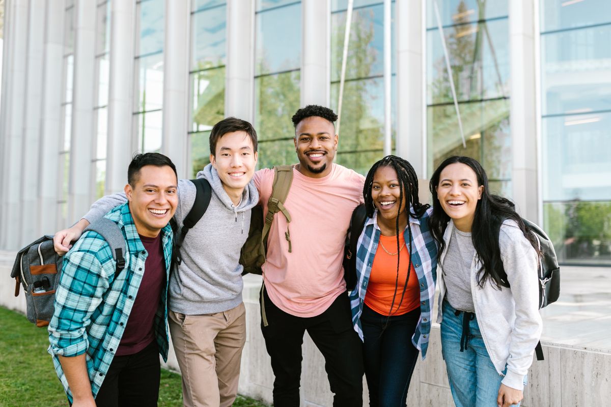 Five people stand together outdoors, smiling and carrying backpacks, perhaps on a break from debating the difference between college and university. A modern glass building rises in the background, embodying the blend of traditional education and contemporary design. Difference Between College and University