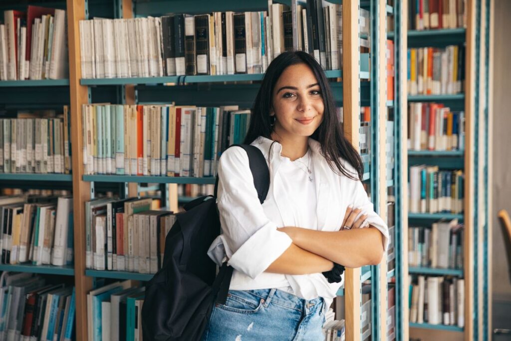 A person with long hair stands smiling in a library, wearing a white shirt and jeans, with a backpack slung over their shoulder, pondering the difference between college and university as they browse the bookshelves. Difference Between College and University.