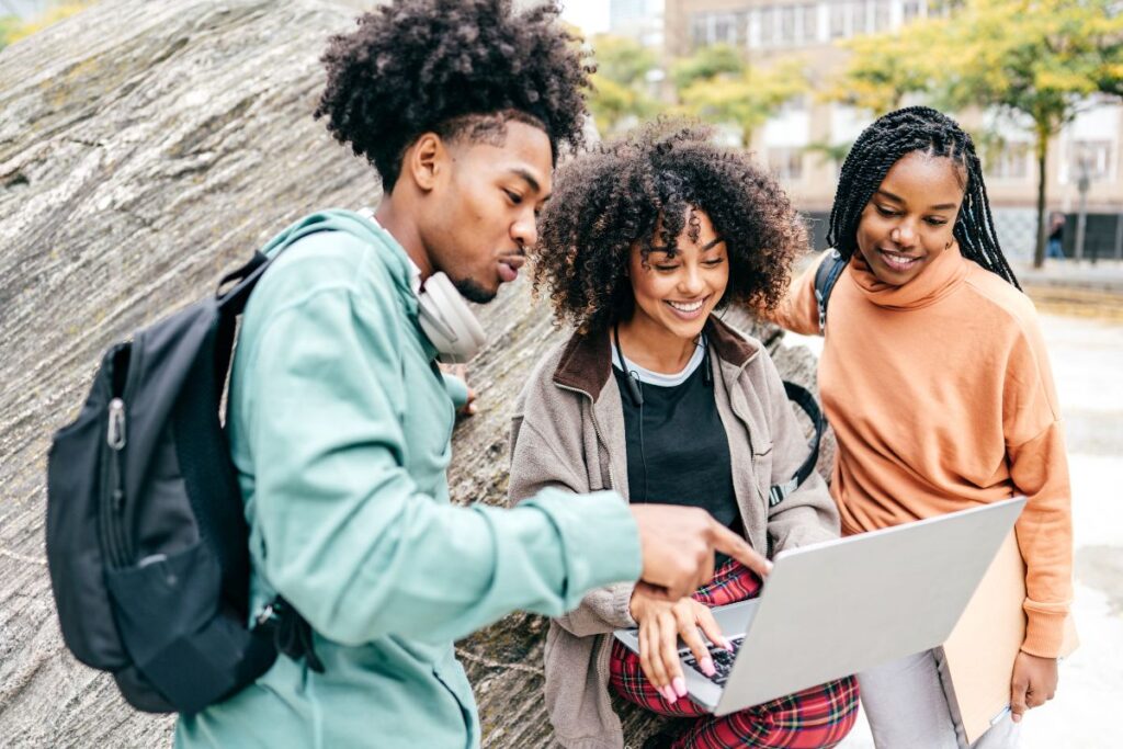 Three people are gathered outdoors, looking at a laptop together. One person points at the screen as they delve into the topic of college vs university. They all appear engaged, sitting near a large rock with buildings in the background.