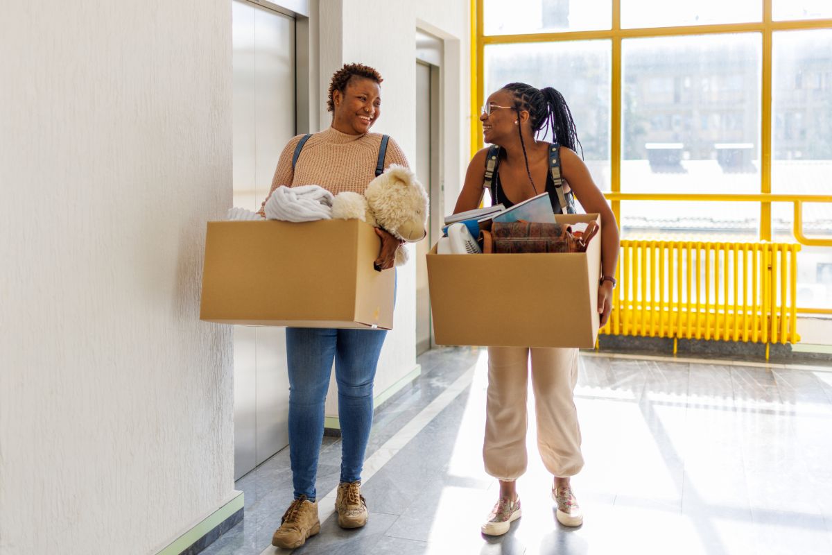 Two people smiling at each other while holding boxes, standing in a brightly lit hallway next to an elevator.