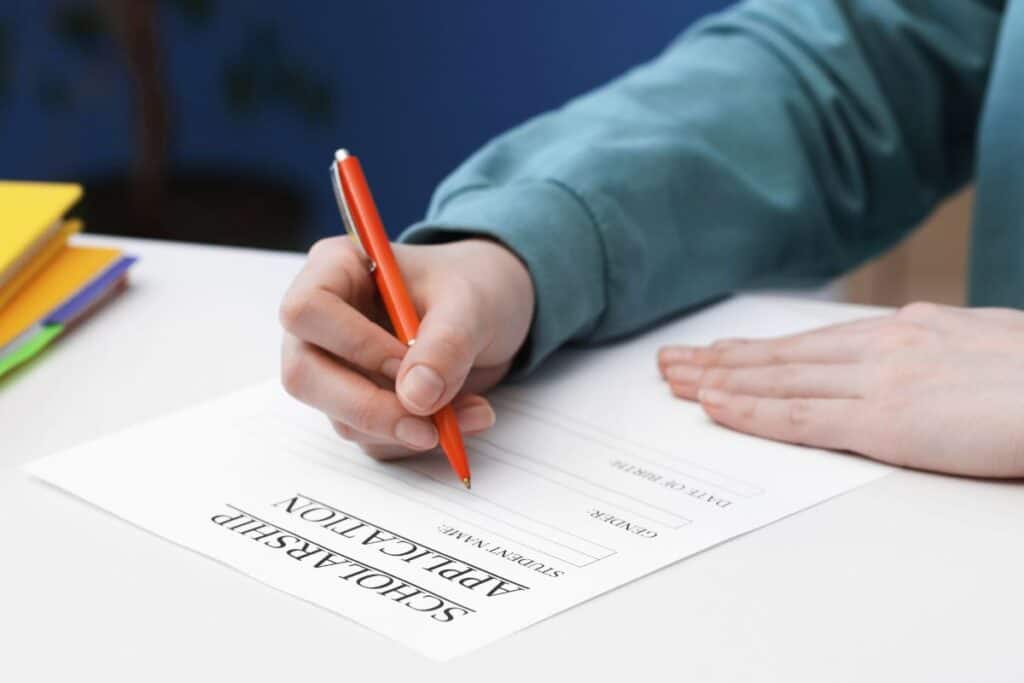 A person fills out a graduate school scholarships application form at a desk, holding an orange pen and wearing a teal long-sleeve shirt.