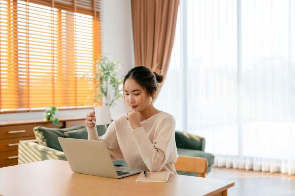 A woman sits at a table with a laptop and notebook, holding a white mug and looking at the screen in a bright, modern living room.