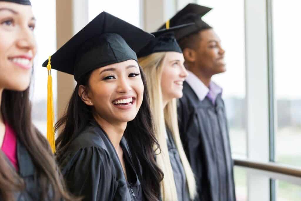Four graduates in caps and gowns stand in a row indoors, smiling and looking toward the right side of the image, celebrating their success with the help of graduate scholarship opportunities.