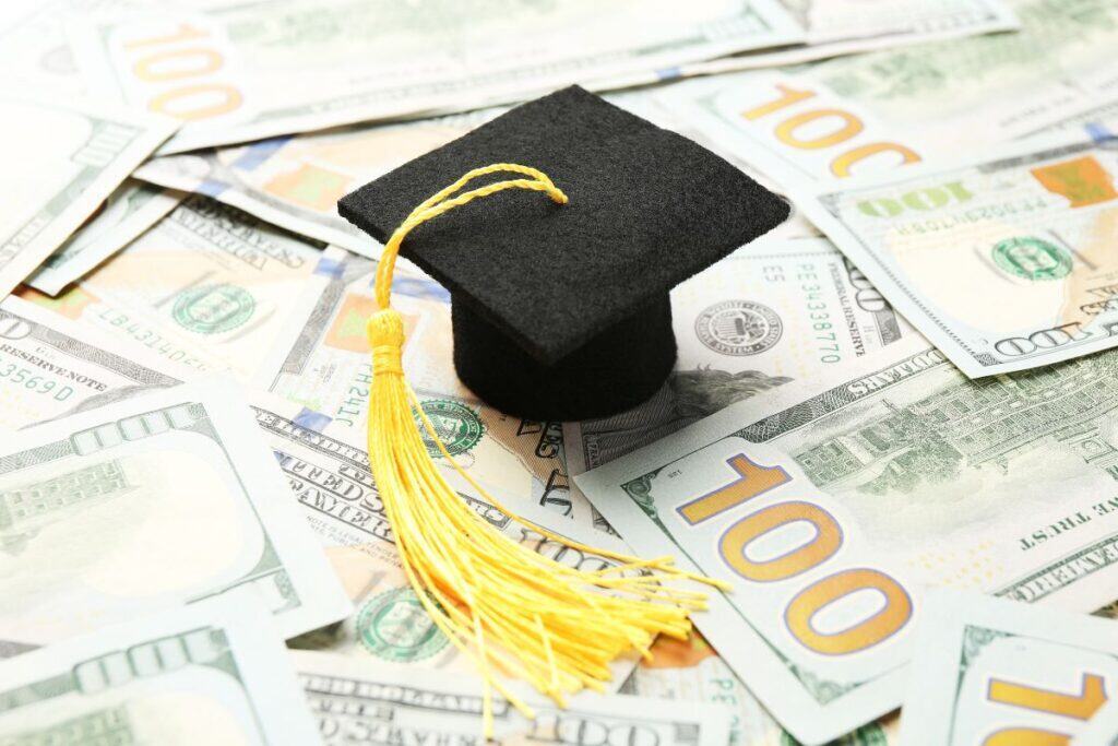 A graduation cap with a yellow tassel rests on a scattered pile of US hundred dollar bills, evoking the question: does FAFSA cover graduate school?.