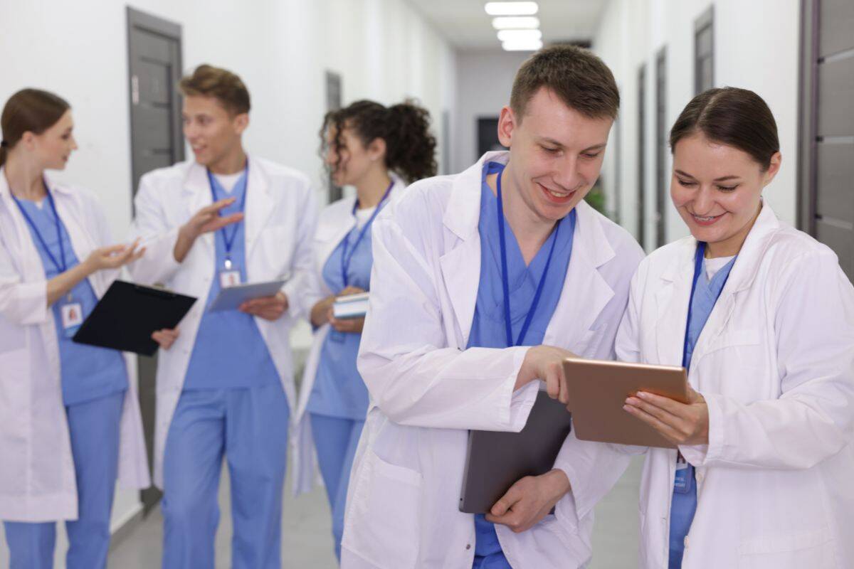 Two medical professionals review information on a tablet in a hallway, while three colleagues converse in the background, all wearing lab coats and scrubs.