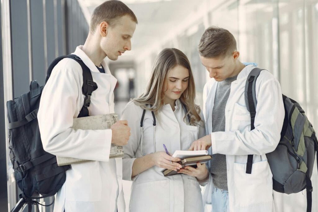 Three medical students in white coats stand together in a hallway, looking at and discussing notes in an open notebook.