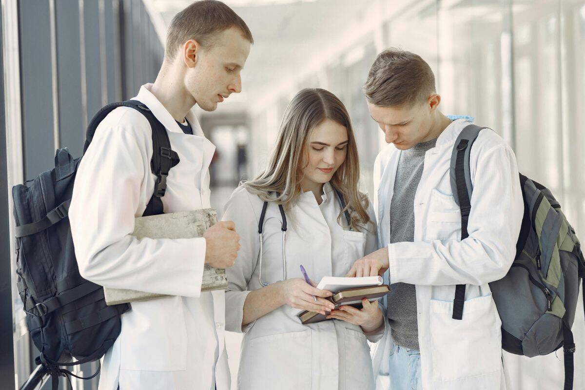 Three medical students in white coats stand together in a hallway, looking at and discussing notes in an open notebook.