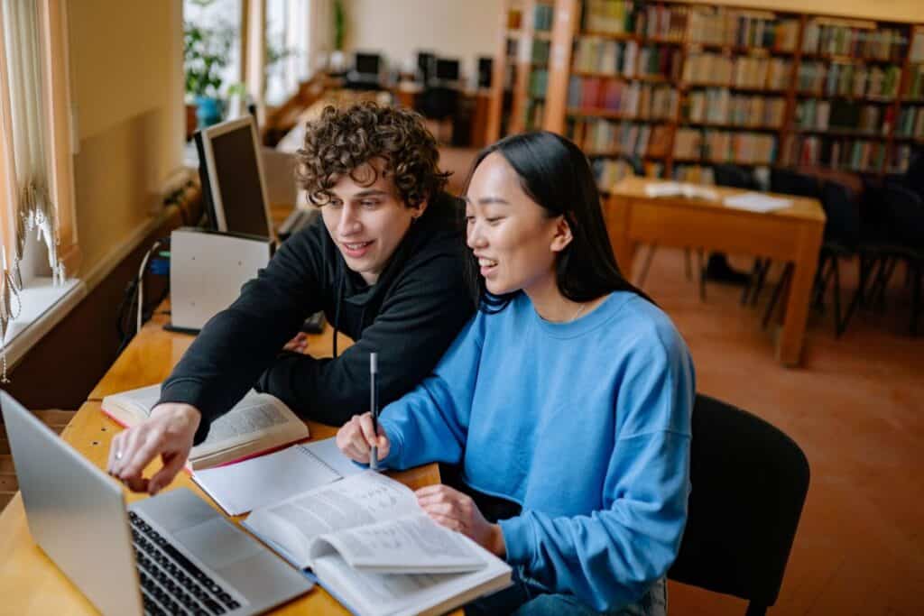 Two students sit at a table in a library, looking at a laptop screen together while studying with open books and notebooks.