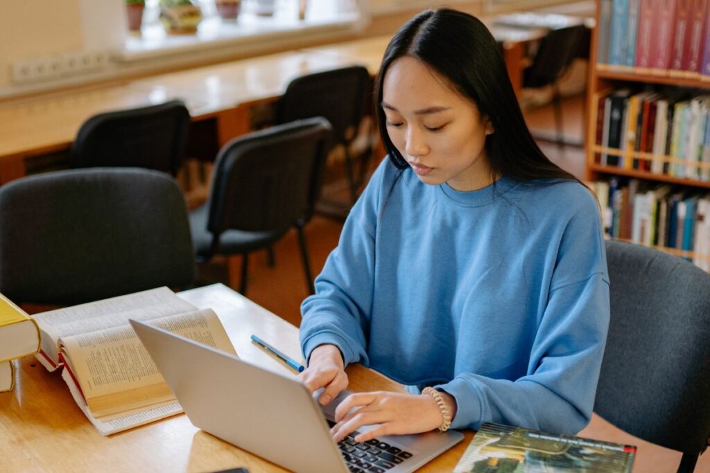 A person wearing a blue sweatshirt types on a laptop at a desk with open books and a pen in a library, possibly drafting medical school personal statement examples.