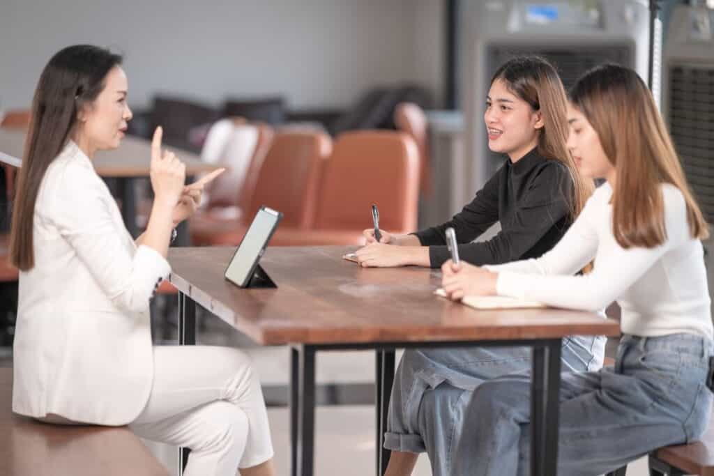 Three women sit at a table in a bright room, with one speaking and gesturing while the other two listen and take notes using notebooks and pens—engaged in an insightful discussion about how to choose a college admissions consultant.