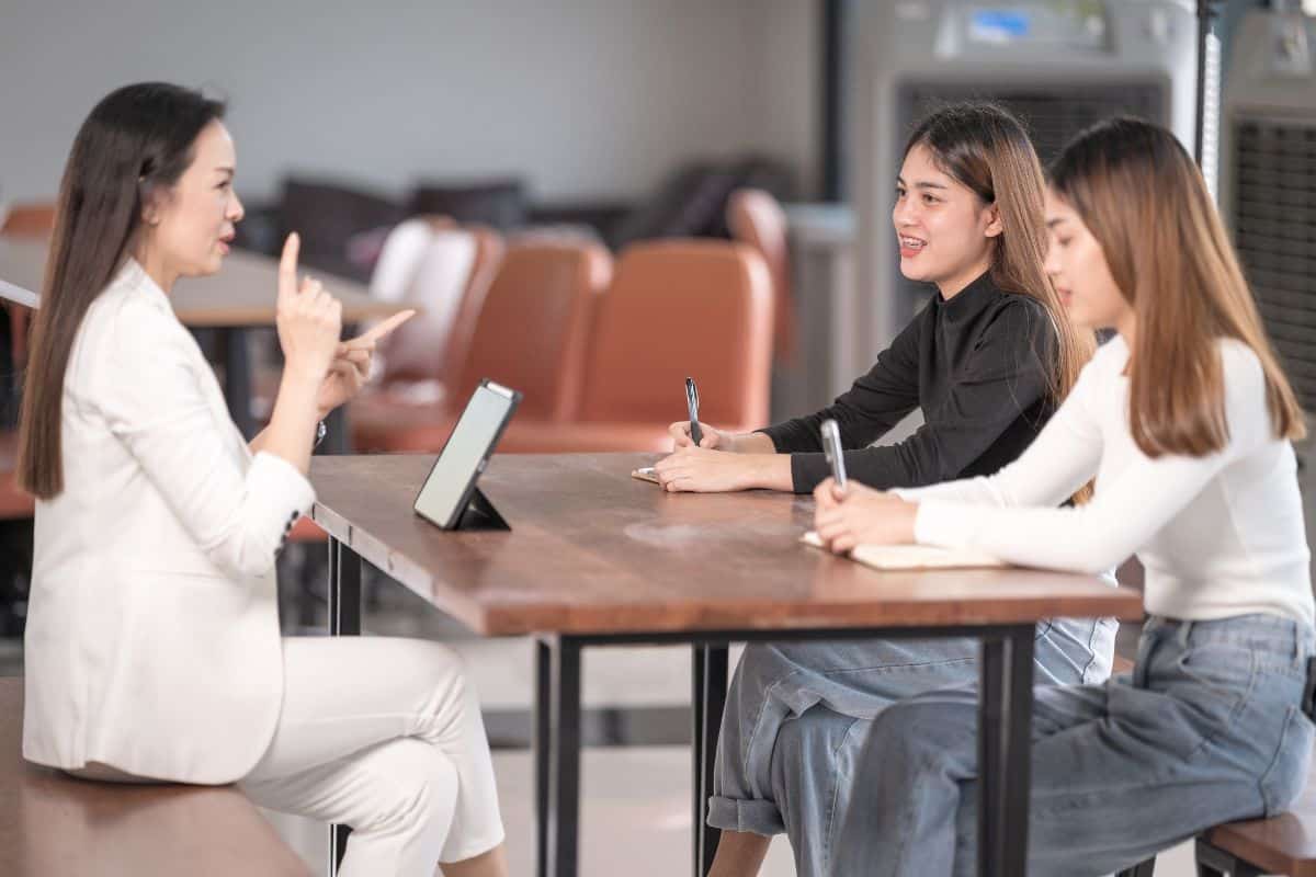 Three women sit at a table in a bright room, with one speaking and gesturing while the other two listen and take notes using notebooks and pens—engaged in an insightful discussion about how to choose a college admissions consultant.