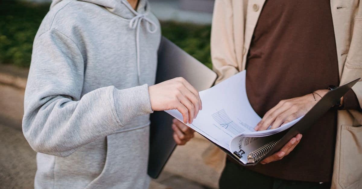 Crop male students in casual wear reading learning material in folder while standing together in university park