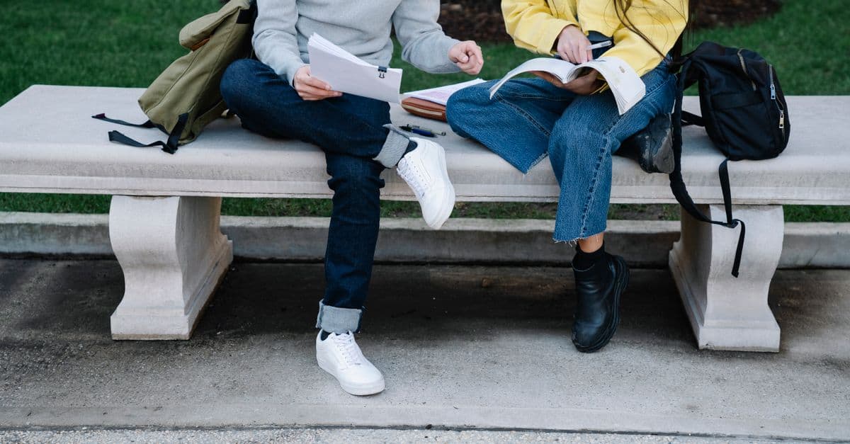 Two students studying outdoors on a concrete bench with books and bags.